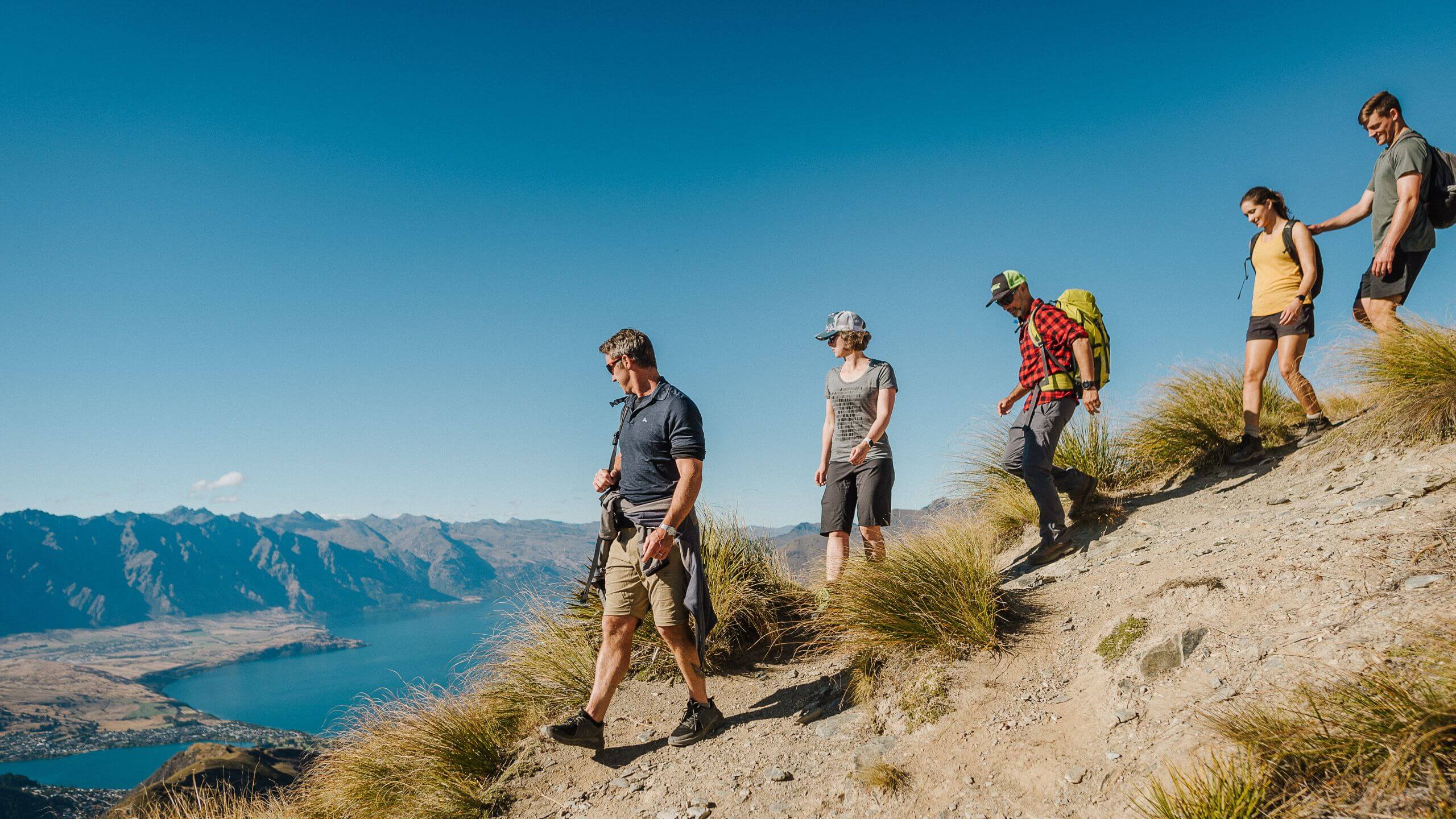 Group of hikers walking down a mountain trail above Queenstown with Lake Wakatipu and The Remarkables in the background.