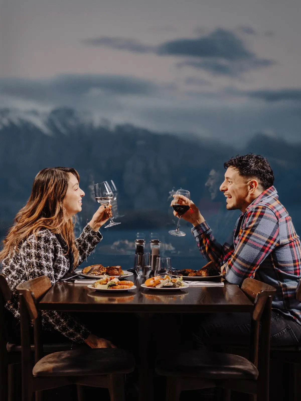 Two diners enjoying a meal and wine at Stratosfare Restaurant in Queenstown, with panoramic lake and mountain views visible through large windows.