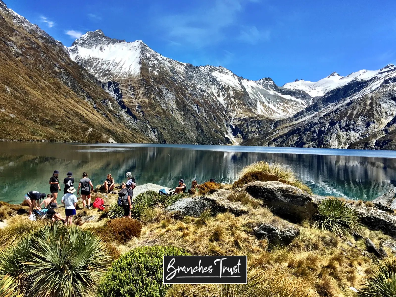 Young people gathered beside an alpine lake surrounded by snow-capped mountains during an outdoor adventure experience supported by Branches Charitable Trust.