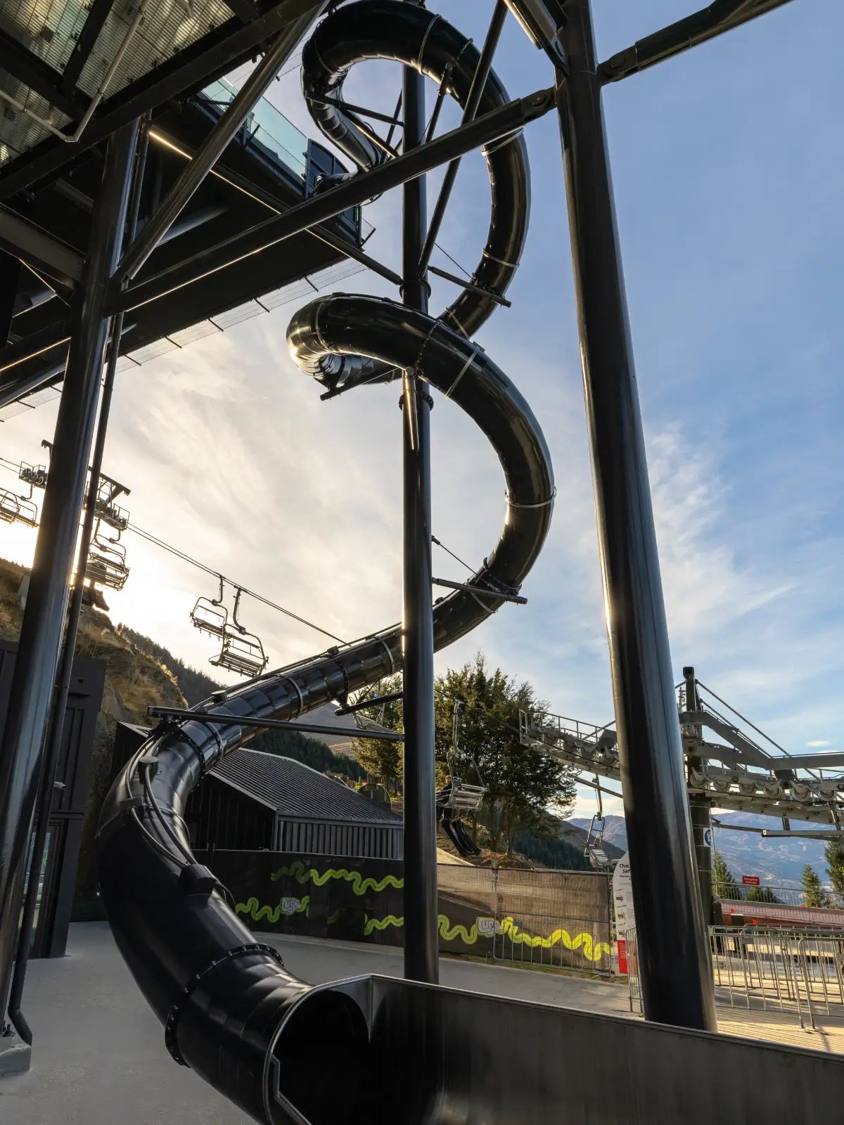 The Vortex Slide at Skyline Queenstown winding through multiple spiral turns beside the gondola terminal, viewed from below against the Queenstown sky.
