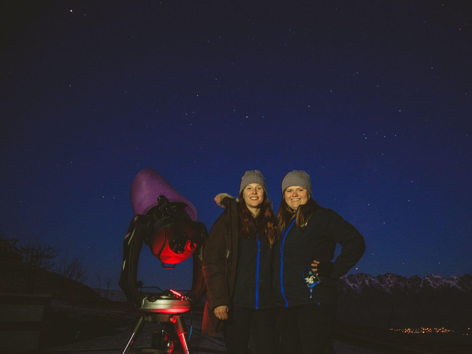 Two people smiling while next to a telescope, with a starry sky in the background