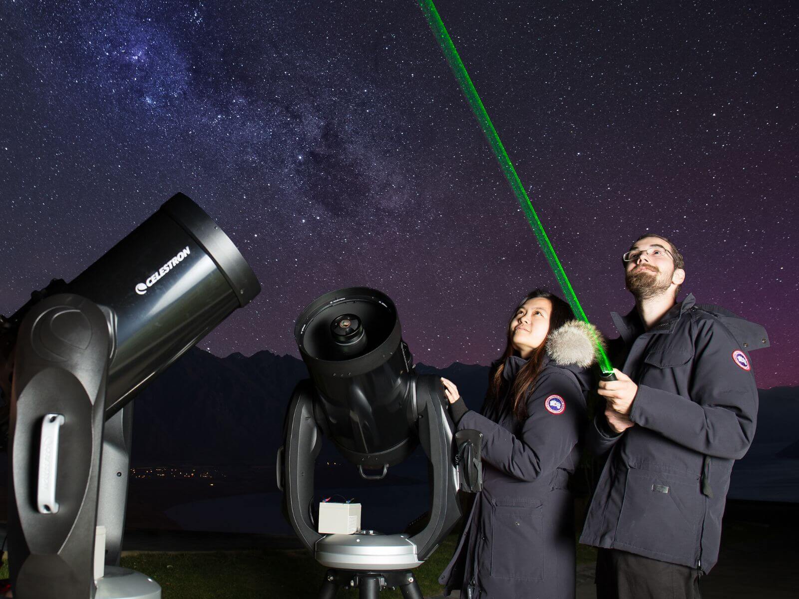 Two people using a laser to point out stars in the background standing next to 2 telescopes